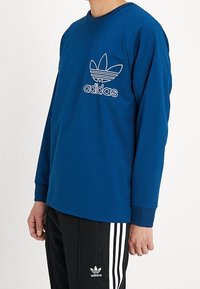Niebieska koszula z długim rękawem, okrągłym dekoltem i białym logo Adidas; noszona z czarnymi spodniami z białymi pionowymi paskami.