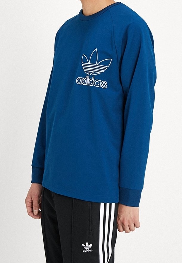 Niebieska koszula z długim rękawem, okrągłym dekoltem i białym logo Adidas; noszona z czarnymi spodniami z białymi pionowymi paskami.