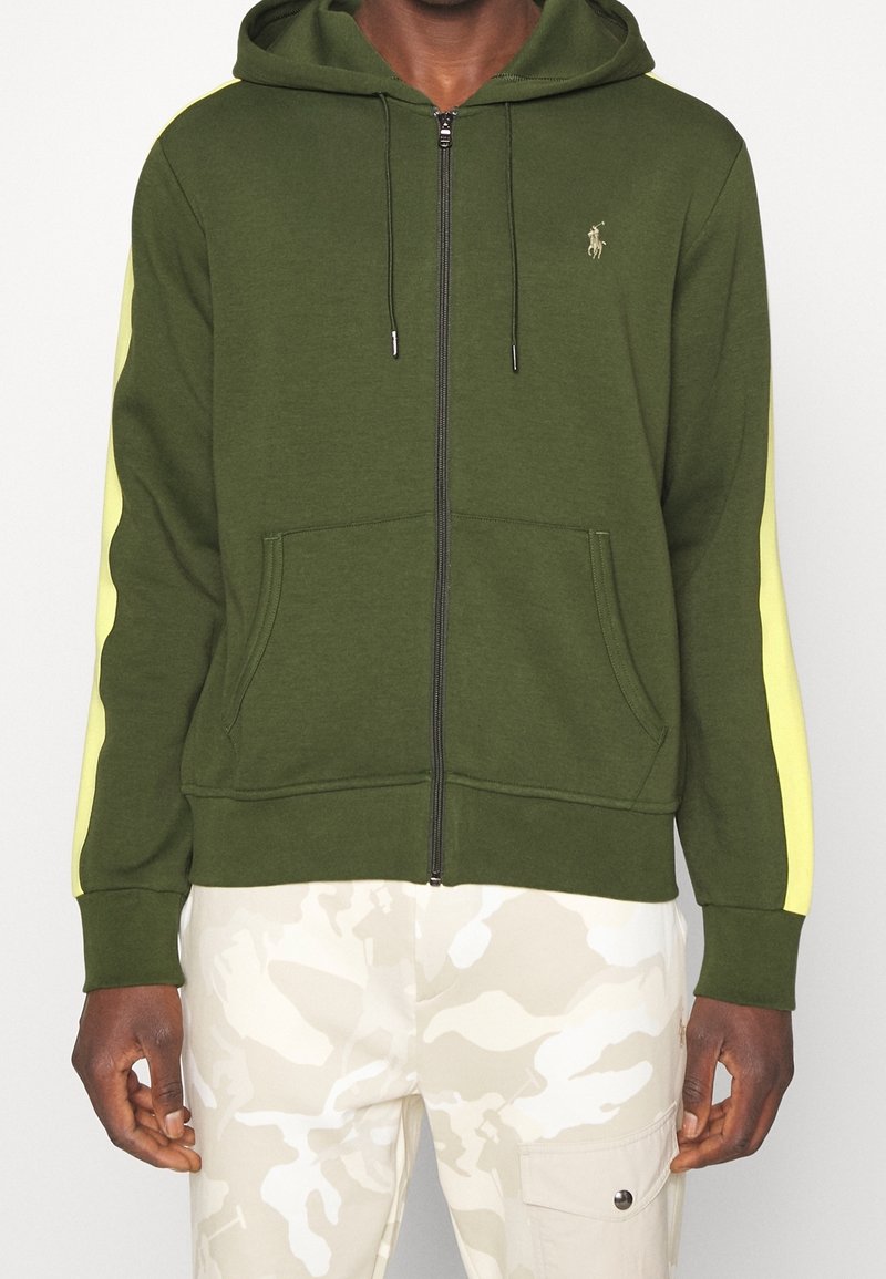 Grön zip-up hoodie med dragsko, med gula sidostrikar och framfickor. Visas med ljus kamouflagemönstrade byxor.