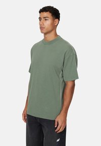 T-shirt verde oversize con maniche corte, colletto a coste e dettagli di cucitura in contrasto. Abbinato a jeans scuri.