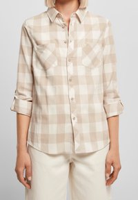 Donna che indossa una camicia a quadri beige e bianca con maniche arrotolate e pantaloni bianchi, in piedi contro uno sfondo grigio chiaro.