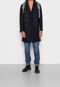 Persona vestida con un abrigo negro de doble botonadura, camisa naranja, jeans azules, botas negras y una mochila gris, de pie contra un fondo plano.