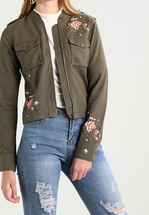 Woman wearing olive green cropped jacket with floral embroidery, white top, and ripped blue jeans, standing against a plain background.