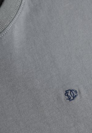 Close-up af gråt stof med syet blåt cirkulært logo nær sømmen på en ribbet krave.