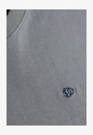 Zbliżenie szarej tkaniny z niebieskim, okrągłym haftowanym logo w pobliżu szwu żeberkowego kołnierza.