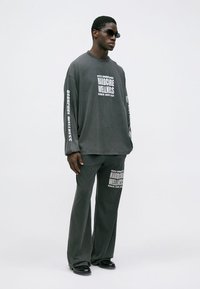 Šedá oversized košeľa s dlhým rukávom a bielym textom, spolu s širokými nohavicami. Text zahŕňa "HARDCORE WELLNESS" a "BERLÍN 2025-2022." Nosia sa slnečné okuliare.
