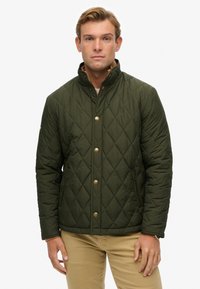 Hombre vistiendo una chaqueta acolchada verde oscura con botones dorados y bolsillos con cremallera, combinada con pantalones beiges, de pie contra un fondo blanco.