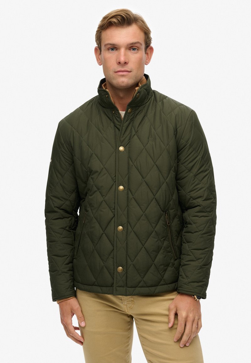 Hombre vistiendo una chaqueta acolchada verde oscura con botones dorados y bolsillos con cremallera, combinada con pantalones beiges, de pie contra un fondo blanco.