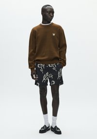 Sweat-shirt marron avec un logo brodé blanc, associé à un short noir présentant un motif abstrait blanc. Chaussettes blanches et chaussures noires.