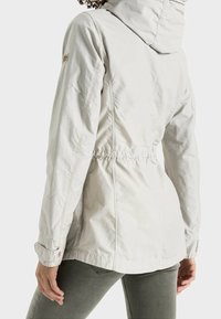 Chaqueta con capucha de color claro, con la cintura ajustada, tejido texturizado y costuras visibles. Presenta un parche con el logotipo en la parte superior del brazo.