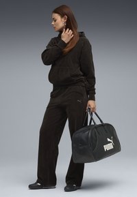 Mørkebrun corduroy hoodie og bukser med frontlomme, parret med en sort læder duffelbag med et hvidt Puma-logo.