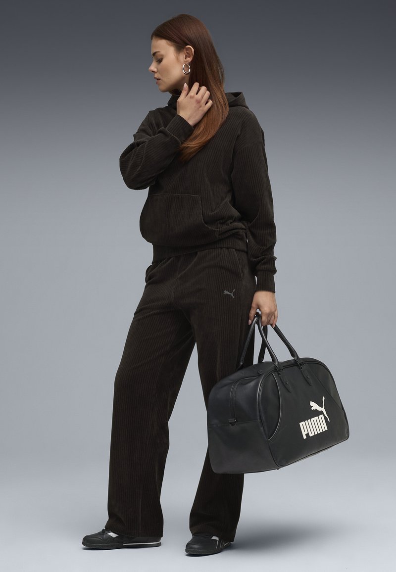 Mørkebrun corduroy hoodie og bukser med frontlomme, parret med en sort læder duffelbag med et hvidt Puma-logo.