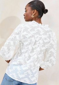 Weißes Blusenshirt mit 3/4-Ärmeln, das ein strukturiertes Muster aus kreisförmigen Applikationen zeigt; Rüschendetails am Ausschnitt; durchsichtiger, leichter Stoff.