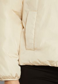 Lys beige puffer-jakke med en glatt, skinnende tekstur, med sidelomme og elastiske mansjetter, designet for en avslappet passform.