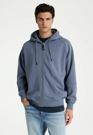 Junger Mann trägt eine blaue Kapuzenjacke mit Reißverschluss, Kapuze und Kordeln, kombiniert mit hellblauen Jeans, steht vor schlichtem Hintergrund.