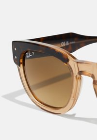 Ochelari de soare cu nuanță maronie, cu o ramă din acetat translucid de culoare bej, având un design îndrăzneț și unghiular. Logo-ul Ray-Ban pe lentilă și pe tâmplă.
