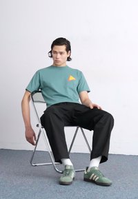 Camiseta teal con un pequeño logo de triángulo naranja, pantalones negros, zapatillas verdes, sentado en una silla de metal contra una pared blanca lisa.
