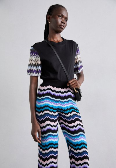 Μαύρη μπλούζα με μοτίβο στις κοντές μωβ, μπλε και μπεζ μανίκες. Παντελόνι με φαρδιά πόδια και ζιγκ ζαγκ ρίγες σε αποχρώσεις του μπλε, μαύρου και ροζ.
