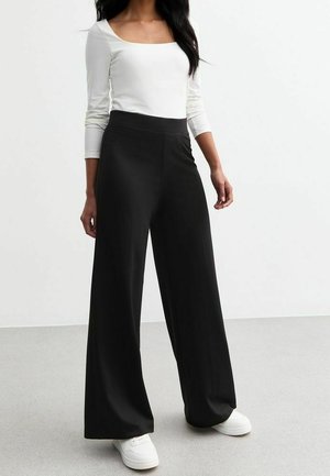 Vrouw die zwarte, wijde broek met hoge taille, een wit shirt met lange mouwen en witte sneakers draagt, staand op een grijze vloer tegen een witte muur.