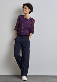 Blusa trasparente a maniche corte con motivo viola e rosso, abbinata a pantaloni a vita alta blu navy e sneakers bianche.