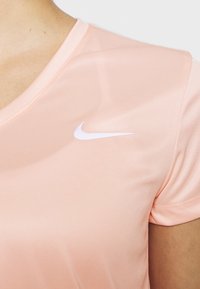 Vaaleanpunainen urheilupaita, joka on valmistettu sileästä kankaasta, ja siinä on pyöreä kaulus sekä pieni valkoinen Nike-logo vasemmassa yläkulmassa.