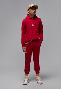 Red sweatshirt and jogger set with black Jordan text, soft fabric, kangaroo pocket, ribbed cuffs, paired with tan cap and sneakers.