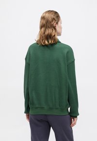 Suéter verde, texturizado, con corte holgado, hombros caídos y puños acanalados. Combinado con pantalones grises, sobre un fondo liso.