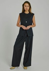 Gilet senza maniche a righe sottili scuro con bottoni frontali, abbinato a pantaloni larghi a righe sottili e tacchi beige appuntiti.