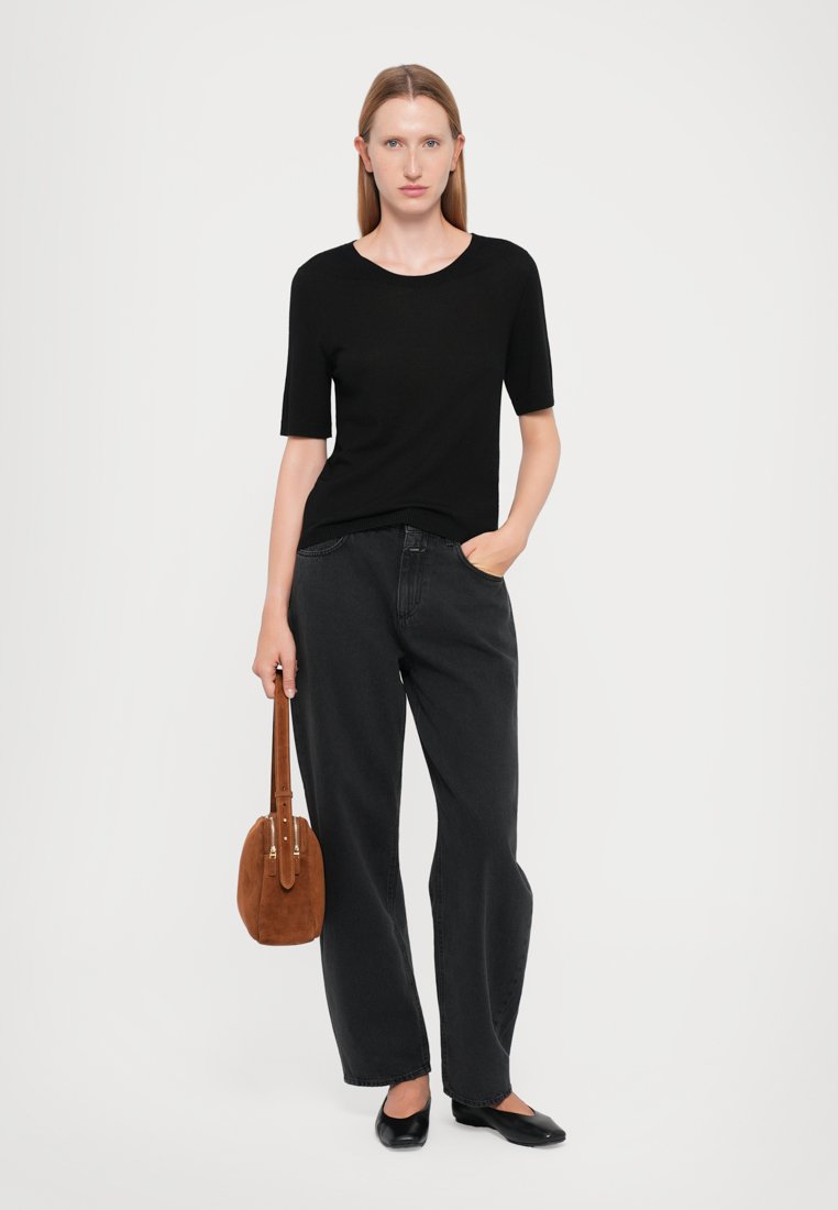 Haut noir à manches courtes associé à un jean noir taille haute. Le mannequin tient un sac en suède beige avec une silhouette courbée et des accessoires en or.