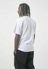 White cotton t-shirt with short sleeves, round neckline, relaxed fit, and smooth texture. Worn with dark pants, showing minimalist design details.