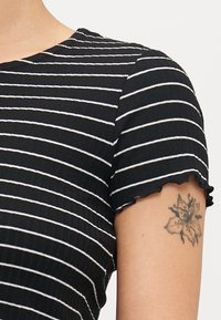 Mustru mitmust ja valged triibud lühikeste varrukatega särk, millel on ribakujuline tekstuur ja lainelised servad, koos lillese tätoveeringuga käel.