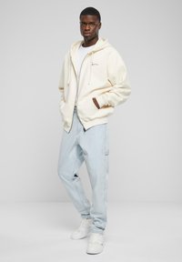 Cream hooded sweatshirt with a zipper, front pockets, and logo detail. Light blue loose-fit jeans and white sneakers.