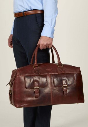 Brown leather duffel bag with two front pockets, buckle accents, and dual handles, held by a person dressed in formal attire.