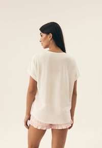 Biała bluzka z krótkim rękawem, wykonana z miękkiego materiału, z podwiniętymi mankietami; zestawiona z różowymi, w kratkę szortami z falbanami. Gładka faktura, luźny krój.