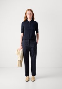 Polo de marină cu mânecă scurtă, cu deschidere cu nasture, combinat cu pantaloni negri bine croiți și pantofi loafers bej. Ținând o jachetă de culoare deschisă.