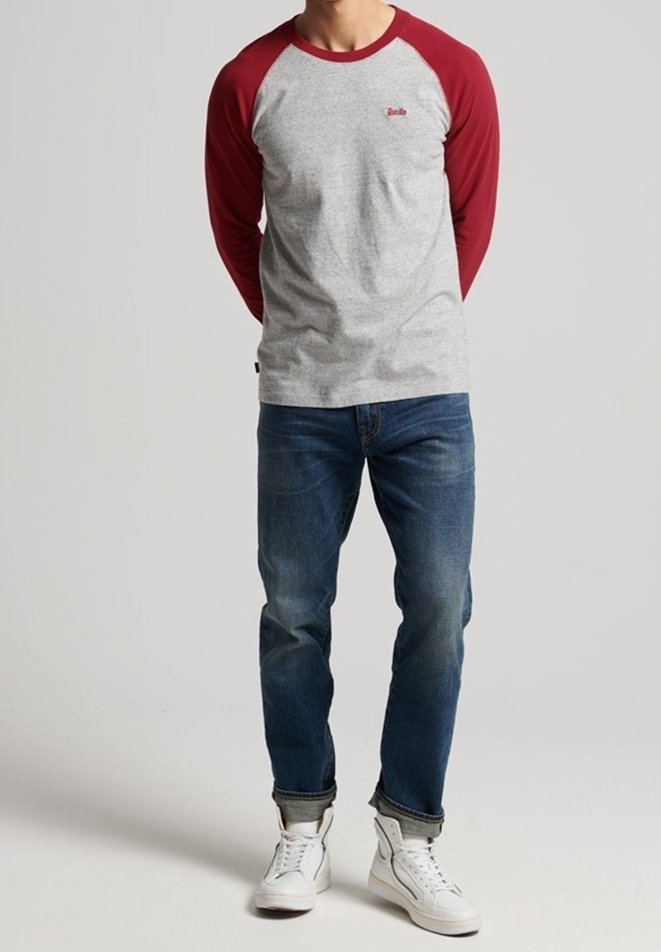 Man med grå och röd långärmad raglantröja, blå jeans med uppvikta ben och vita hög-top sneakers, stående mot en enfärgad bakgrund.