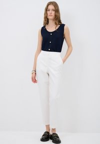 Marineblauwe geribbelde tanktop met gouden knopen, gecombineerd met witte op maat gemaakte broek en zwarte instappers. Gladde texturen, hedendaagse snit.