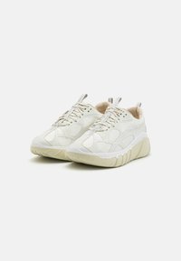 Valkoiset tennarit, jotka on tehty teksturoidusta synteettisestä materiaalista, paksulla vaaleanharmaalla pohjalla. Ominaisuuksiin kuuluvat verkkomaiset yksityiskohdat ja huomaamaton brändäys sivussa.