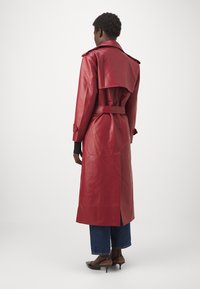 Rød læder trench coat med bælte i taljen, bred krave og slidser i siderne, parret med blå denim jeans og sorte højhælede sko.