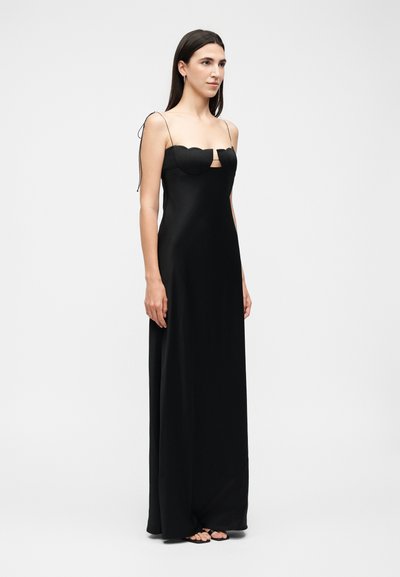 Γυναίκα που φορά μαύρο μακρύ φόρεμα με κυματιστό ντεκολτέ και λεπτές τιράντες, φοράει μαύρα σανδάλια με λευκό φόντο.