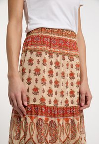 Maxi-jurk van zachte stof met rode bloempatronen op een beige achtergrond. Voorzien van een elastische tailleband en een gelaagd ontwerp met contrasterende accenten.