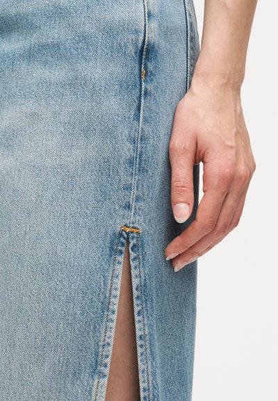 Δεξί χέρι με φυσικά νύχια ακουμπισμένο δίπλα σε φούστα από ανοιχτό μπλε τζιν με εμπρός σκίσιμο και εμφανείς ραφές.