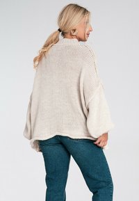 Sweter z dzianiny w jasnym beżu, oversize'owy krój, luźne rękawy, faktura prążkowana, okrągły dół, noszony z niebieskimi dżinsami z wysokim stanem.