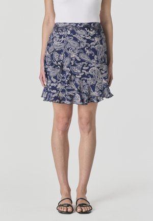 Mujer vistiendo una falda azul marino con patrones de cachemira blancos y dobladillo con volantes, combinada con una blusa blanca y sandalias negras.