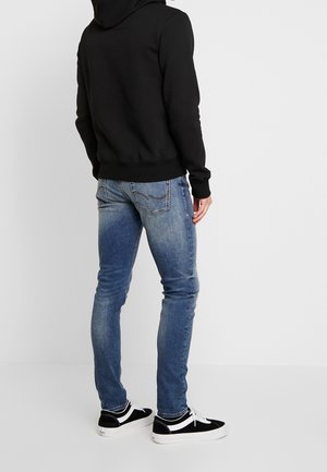 Zwarte hoodie, slim-fit blauwe jeans, zwart-witte sneakers. De hoodie heeft een capuchon en geribbelde manchetten; de jeans hebben een vervaagde textuur met zakken.