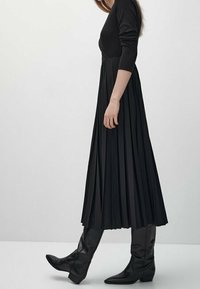 Robe noire plissée à manches longues, en tissu léger, associée à des bottes noires montantes, mettant en valeur une silhouette épurée.