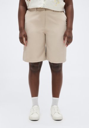 Person med mørk hud som har på seg beige shorts til knærne, hvite sokker, hvite joggesko og en lysfarget, blomstrete ermeløs topp.