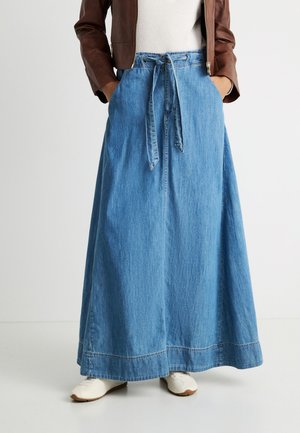 Persoon die een lichtblauwe denim maxi-rok draagt met een vooraan knoopceintuur, witte sneakers, een wit topje en een bruine leren jas, met de handen in de zakken.