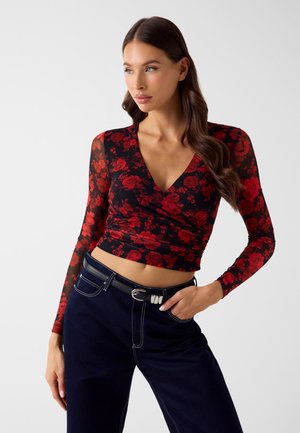 Czerwona bluzka w kwiaty z długimi przezroczystymi rękawami i głębokim dekoltem w kształcie litery V, zestawiona z ciemnymi dżinsami i czarnym paskiem. Gładka faktura i dopasowany krój.