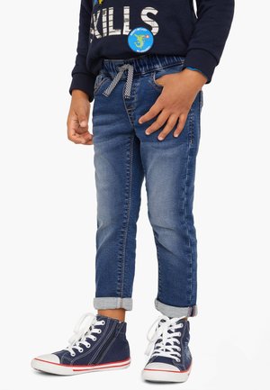 Child wearing rolled-up blue jeans, navy high-top sneakers with white laces, and a navy sweatshirt with patches and text.